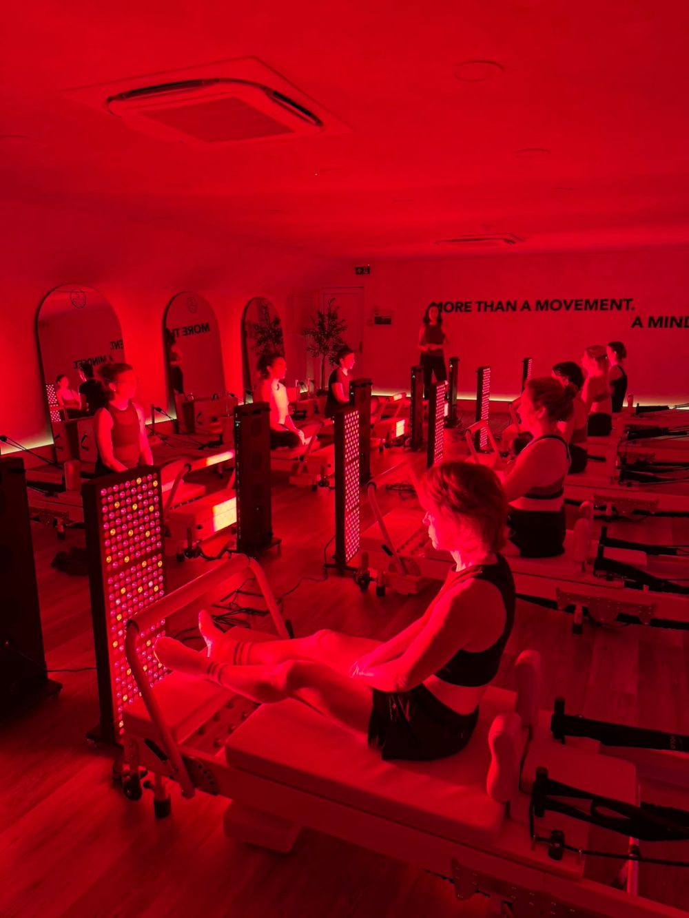 Reformer Pilates class in a red-lit studio with a bodybud red light panel beside the beds